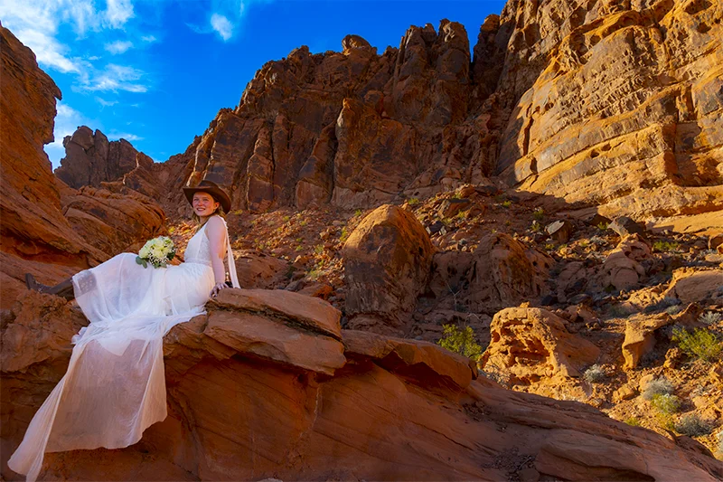 Smiling bride in flowing white dress and cowboy hat seated on red sandstone rocks holding a bouquet at Valley of Fire