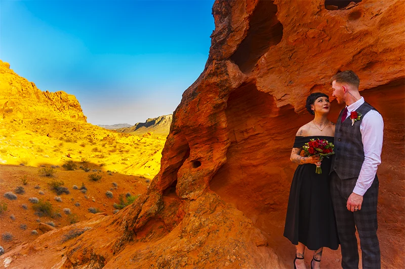 Couple in black wedding attire with red rose bouquet standing inside a curved sandstone rock alcove at Valley of Fire