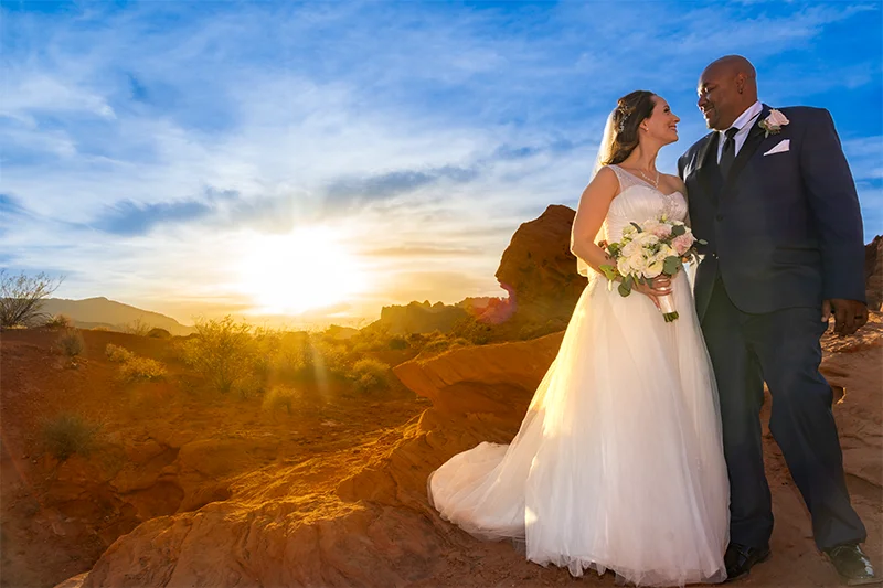 Bride in white ball gown and groom in dark suit looking at each other against a dramatic golden sunset and red rock formations at Valley of Fire