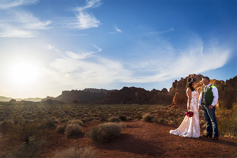 Bride in white lace gown and groom in green vest sharing a sunset moment on the open desert floor at Valley of Fire