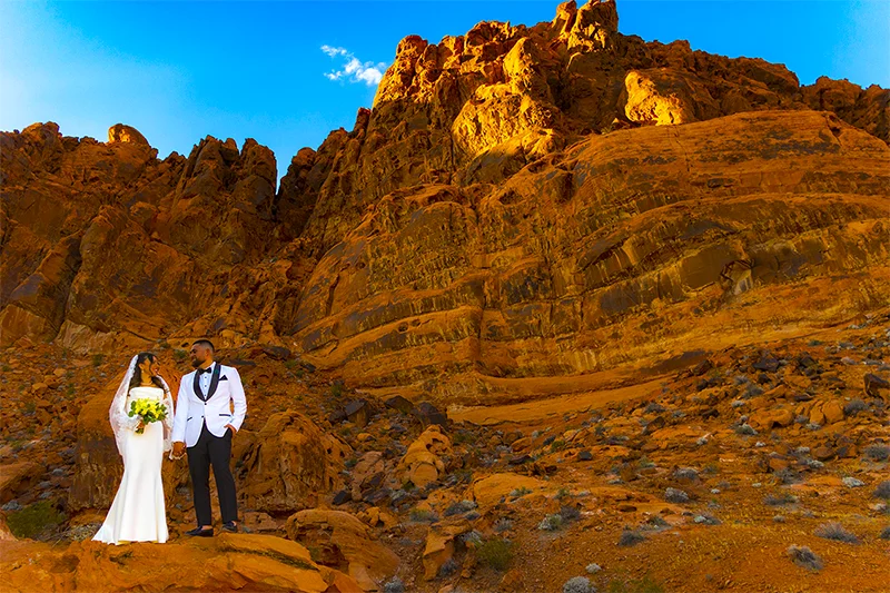 Bride in white gown and groom in dark suit standing against towering red sandstone cliffs at Valley of Fire